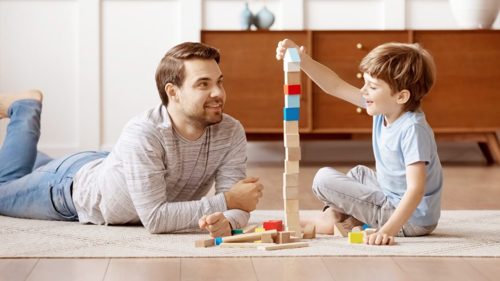 Mann baut mit Kind einen Turm aus Holzbausteinen am Boden